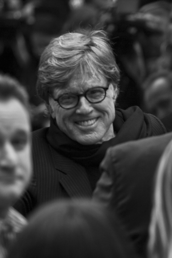 Robert Redford smiling in a vintage film set photo, behind-the-scenes with a director’s chair and clapperboard in view.
